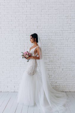 Beyaz elbiseli ve peçeli güzel gelinin portresi beyaz tuğla duvarda çiçek demeti ile poz veriyor.             