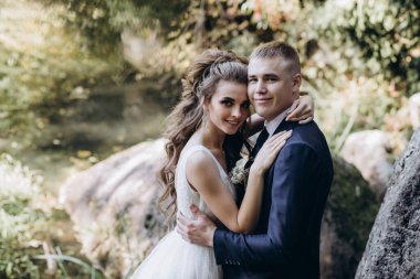 Genç bir çift düğün günlerinde güneşli bir yaz parkında romantik bir anın tadını çıkarır. Doğayla çevrili, gülümsüyor ve birbirlerine sevgiyle sarılıyorlar..