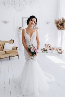 Beyaz elbiseli güzel genç gelin zarif iç kısımda düğün buketi ile poz veriyor.          
