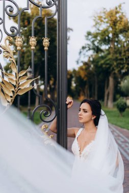 Beyaz gelinlik ve duvak içinde genç ve güzel bir gelin parkın kapısında poz veriyor.       
