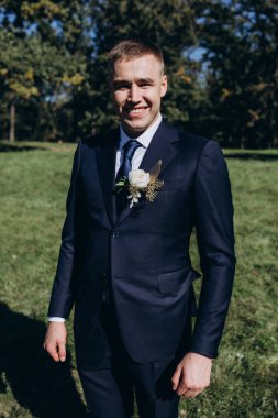 Gelinlik giymiş neşeli genç damat parkta kameraya gülümsüyor.  