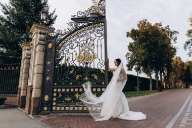 Beyaz gelinlik ve duvak içinde genç ve güzel bir gelin parkın kapısında poz veriyor.       