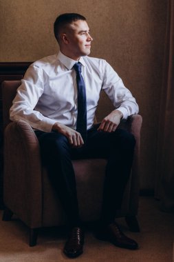 Odada oturan takım elbiseli ve kravatlı yakışıklı genç adam.