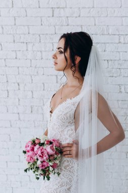 Beyaz elbiseli ve peçeli güzel gelinin portresi beyaz tuğla duvarda çiçek demeti ile poz veriyor.             