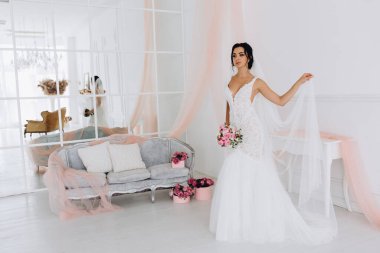 Beyaz elbiseli güzel genç gelin zarif iç kısımda düğün buketi ile poz veriyor.          