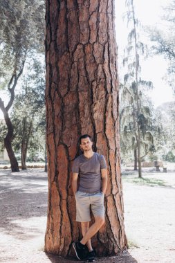 Genç adam parkta bir ağaç gövdesine yaslanıyor.