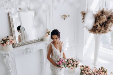 Beyaz elbiseli güzel genç gelin zarif iç kısımda düğün buketi ile poz veriyor.          