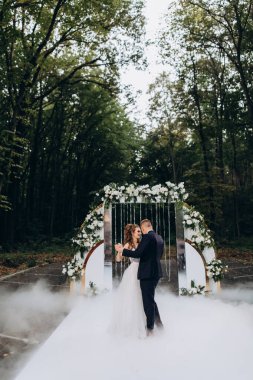 Güzel, mutlu, genç bir çift. Dekoratif çiçek kemerinin yanında dans ediyorlar.          