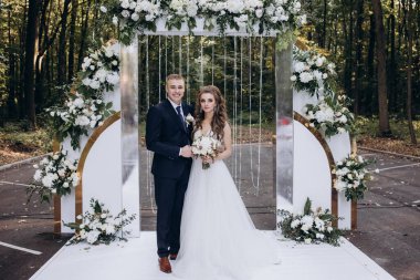 Mutlu gelin ve damat parkta düğün kemerinin yanında poz veriyor.          