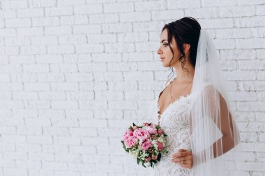 Beyaz elbiseli ve peçeli güzel gelinin portresi beyaz tuğla duvarda çiçek demeti ile poz veriyor.             