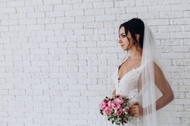 Beyaz elbiseli ve peçeli güzel gelinin portresi beyaz tuğla duvarda çiçek demeti ile poz veriyor.             