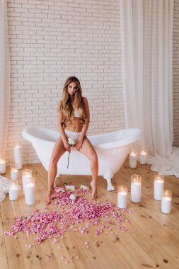 Çiçekler ve mumlarla banyoda poz veren güzel genç bir kadın.        
