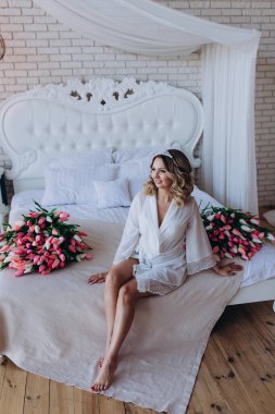 Beyaz bornozlu genç ve güzel gelin yatağında çiçeklerle oturuyor..