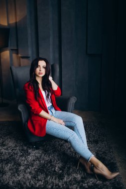 Sandalyede oturan kırmızı ceketli ve kot pantolonlu genç kadın.