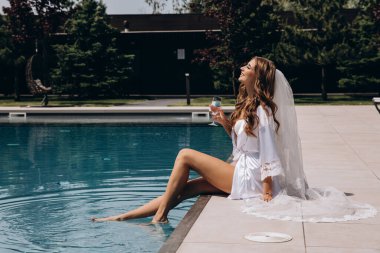 Beyaz peignoir giymiş güzel gelin şarap kadehiyle havuz kenarında poz veriyor.
