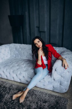 Genç, güzel, esmer, kırmızı ceketli kadın kanepede oturuyor. 