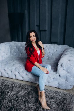 Genç, güzel, esmer, kırmızı ceketli kadın kanepede oturuyor. 