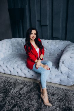 Genç, güzel, esmer, kırmızı ceketli kadın kanepede oturuyor. 