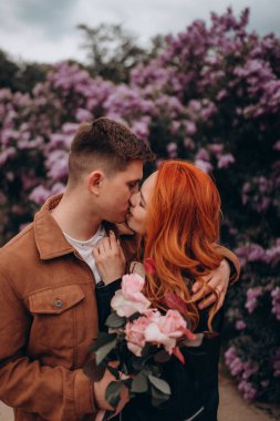 Parkta çiçek açan ağaçlarla sarılıp öpüşen genç bir çift. 