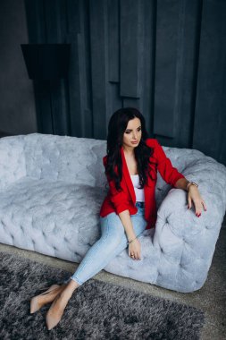 Genç, güzel, esmer, kırmızı ceketli kadın kanepede oturuyor. 