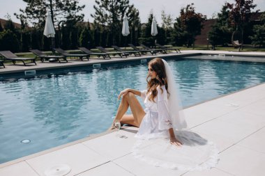 Beyaz peignoir giymiş güzel gelin şarap kadehiyle havuz kenarında poz veriyor.