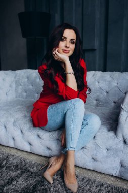 Genç, güzel, esmer, kırmızı ceketli kadın kanepede oturuyor. 