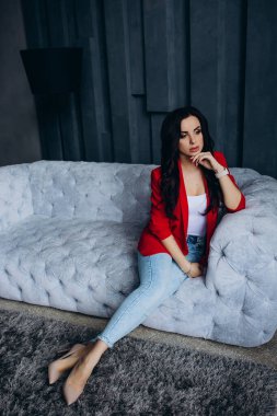 Genç, güzel, esmer, kırmızı ceketli kadın kanepede oturuyor. 