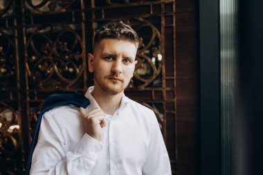 Beyaz tişörtlü, takım elbiseli genç ve yakışıklı bir adam kameraya bakıyor.