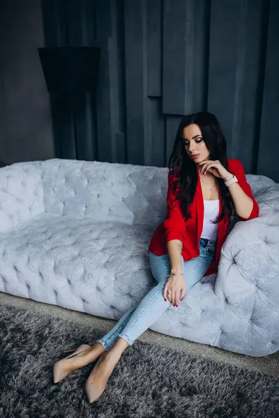 Genç, güzel, esmer, kırmızı ceketli kadın kanepede oturuyor. 