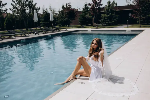 Beyaz peignoir giymiş güzel gelin şarap kadehiyle havuz kenarında poz veriyor.