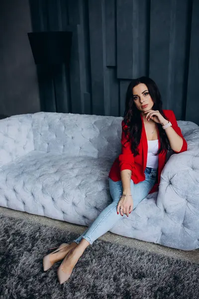 Genç, güzel, esmer, kırmızı ceketli kadın kanepede oturuyor. 