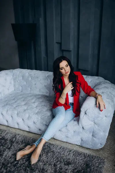 Genç, güzel, esmer, kırmızı ceketli kadın kanepede oturuyor. 