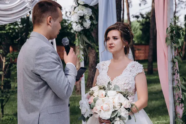 Gelin ve buketi düğün töreninde mikrofonlu yakışıklı damada bakıyor.