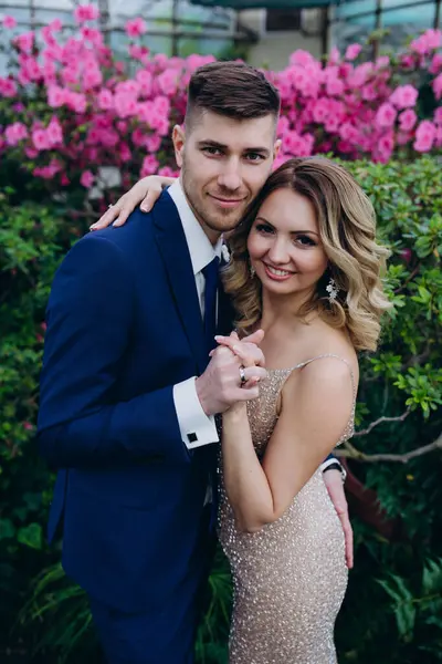 Mutlu gelin ve damat, botanik bahçesinde mor çiçeklerin önünde köprüde kucaklaşıyorlar. 