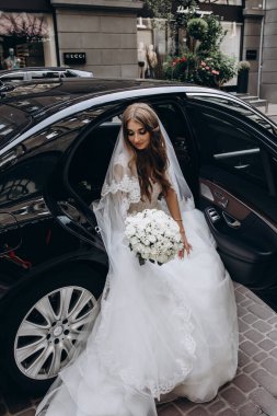 Şık beyaz gelinlikli gelin sokakta arabada poz veriyor.