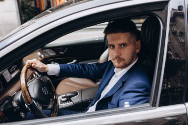 Takım elbiseli genç yakışıklı damat şehir sokaklarında arabada poz veriyor.