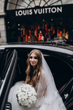 Şık beyaz gelinlikli gelin sokakta arabada poz veriyor.