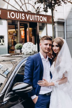 Gelin ve damat düğün gününde şehirde arabanın yanında poz veriyorlar..