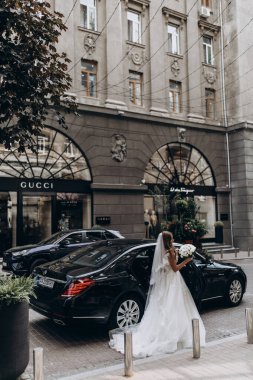 Şık beyaz gelinlikli gelin sokakta arabanın yanında poz veriyor.