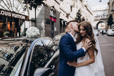 Gelin ve damat düğün gününde şehirde arabanın yanında poz veriyorlar..