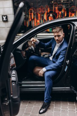 Takım elbiseli genç yakışıklı damat şehir sokaklarında arabada poz veriyor.