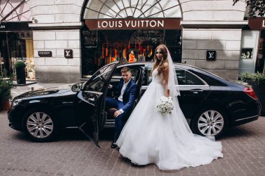 Gelin ve damat düğün gününde şehirde arabanın yanında poz veriyorlar..