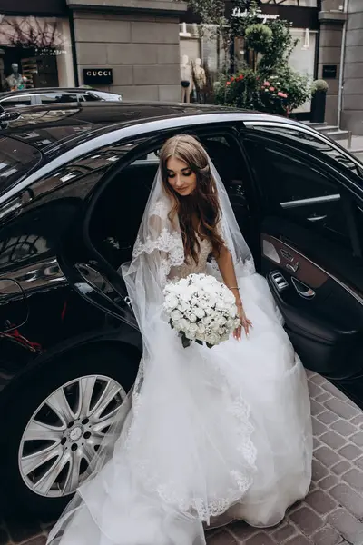 Şık beyaz gelinlikli gelin sokakta arabada poz veriyor.