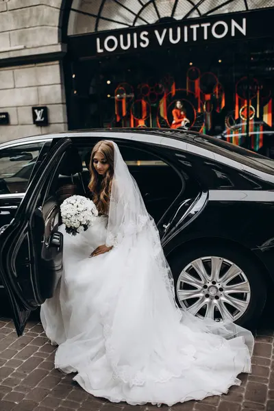 Şık beyaz gelinlikli gelin sokakta arabada poz veriyor.