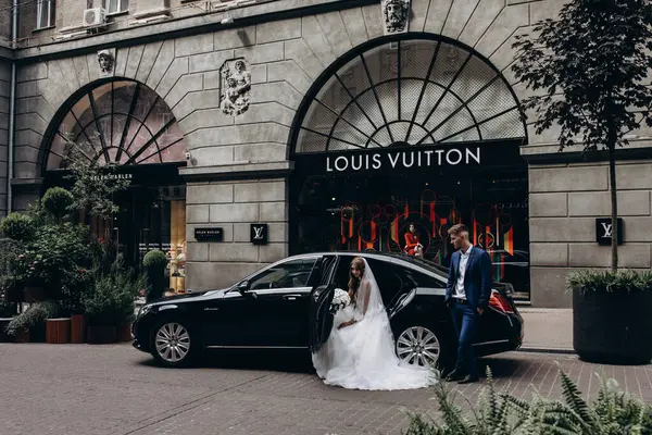 Gelin ve damat düğün gününde şehirde arabanın yanında poz veriyorlar..