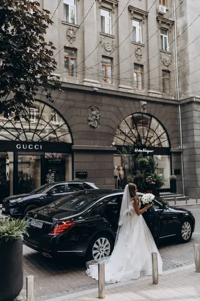 Şık beyaz gelinlikli gelin sokakta arabanın yanında poz veriyor.