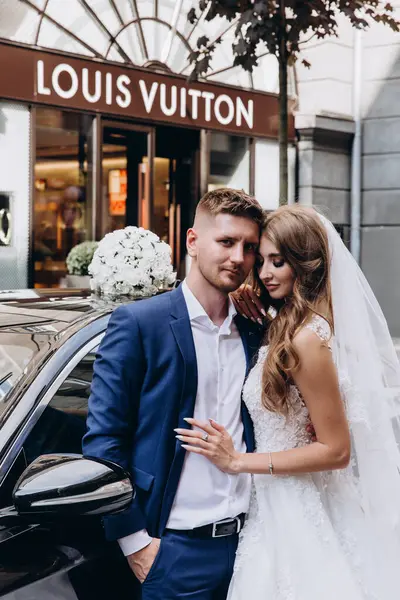 Gelin ve damat düğün gününde şehirde arabanın yanında poz veriyorlar..