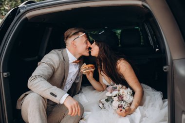 Gelin şapkalı ve damadı arabanın bagajında oturmuş hamburger yiyor ve öpüşüyor.  