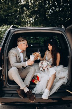 Gelin ve damat bir arabanın bagajında oturmuş biraz yemek yiyor, kola içiyor ve hamburger yiyor. 