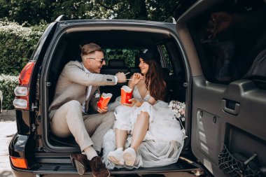 Gelin şapkalı ve damadı arabanın bagajında oturmuş patates kızartması yiyor. 